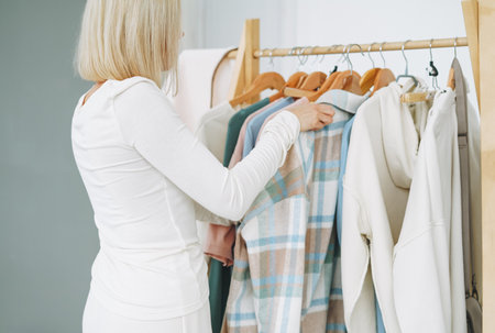 Young blonde woman stylist seller chooses clothes on hanger in bright studioの写真素材