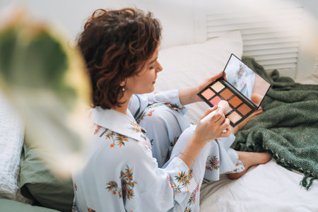 Young brunette woman in blue pajamas applies makeup with face sculpting palette sitting on bed at homeの写真素材