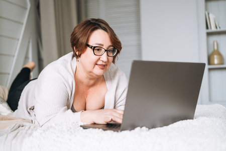 Smiling middle aged plus size woman lying on bed, working on laptop from homeの写真素材