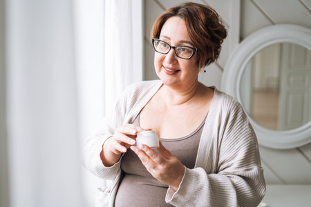 Smiling middle aged plus size woman with day cream near window at homeの写真素材
