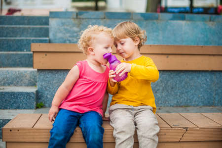 Toddler boy and girl sitting on a bench and drinking waterの写真素材