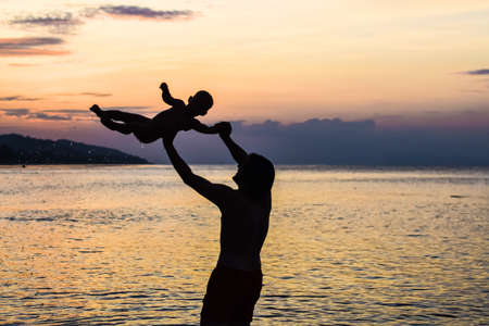 young happy father holding up in his arms little son putting him up at the beach in barefoot standing in front of sea waves wet sand having fun with the kid in Summer sunset coast.の写真素材