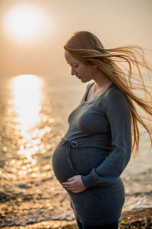 Pregnant young woman standing on the beach in a gray knitted dress, sunset, Pregnancy conceptの写真素材