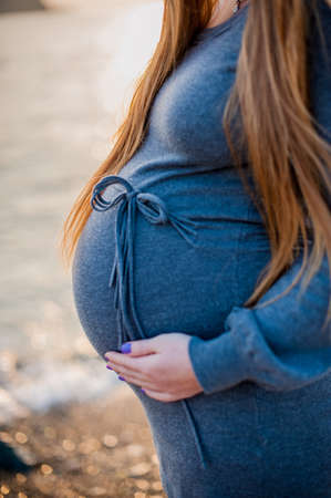 Pregnant young woman standing on the beach in a gray knitted dress, sunset, Pregnancy conceptの写真素材