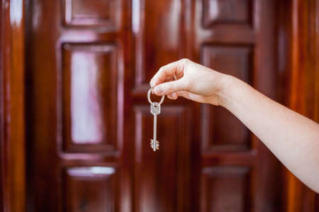 A female hand holding a key to the house on a background of a wooden door. Owning real estate concept.の写真素材
