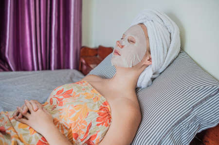 Young red-haired woman relaxing on a bed. Sheet mask on her face. Beauty and Skin Care Conceptの写真素材