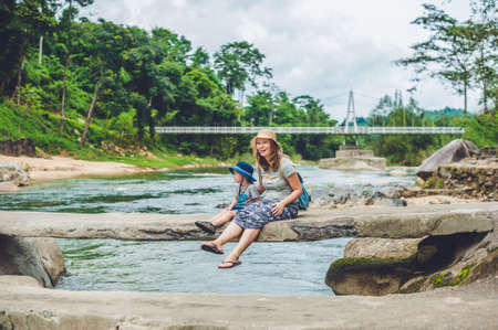 Mom and young son sitting on bridge on a background of the river. Travelling with kids conceptの写真素材