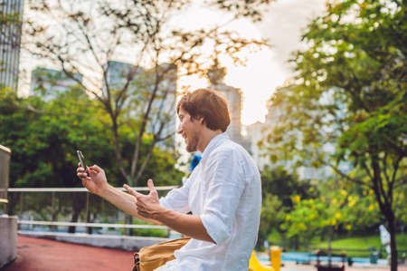 Young Man on the phone. Dressing in a white shirt, beige shorts. Young handsome businessman in casual clothes is sitting outside a business district, making a call on a mobile phone. Mobile Office concept.の写真素材