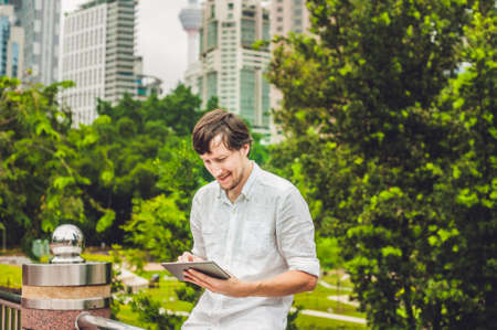 Man businessman or student in casual dress using tablet in a tropical park on the background of skyscrapers. Dressing in a white shirt, beige shorts. Young handsome businessman in casual clothes is sitting outside a business district. Mobile Office concepの写真素材