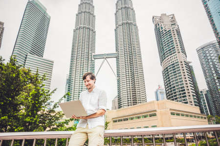 Man businessman or student in casual dress using laptop in a tropical park on the background of skyscrapers. Dressing in a white shirt, beige shorts. Young handsome businessman in casual clothes is sitting outside a business district. Mobile Office concepの写真素材