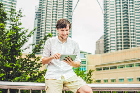 Man businessman or student in casual dress using tablet in a tropical park on the background of skyscrapers. Dressing in a white shirt, beige shorts. Young handsome businessman in casual clothes is sitting outside a business district. Mobile Office concepの写真素材