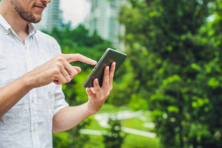 Man businessman or student in casual dress using tablet in a tropical park on the background of skyscrapers. Dressing in a white shirt, beige shorts. Young handsome businessman in casual clothes is sitting outside a business district. Mobile Office concepの写真素材
