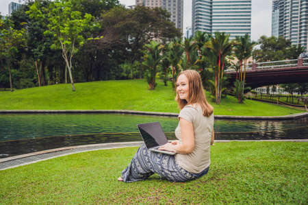 A young woman in casual dress using laptop in a tropical park on the background of skyscrapers. Mobile Office concept.の写真素材