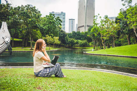 A young woman in casual dress using laptop in a tropical park on the background of skyscrapers....