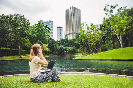A young woman in casual dress using laptop in a tropical park on the background of skyscrapers. Mobile Office concept.の写真素材