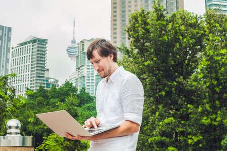 Man businessman or student in casual dress using laptop in a tropical park on the background of skyscrapers. Dressing in a white shirt, beige shorts. Young handsome businessman in casual clothes is sitting outside a business district. Mobile Office concepの写真素材