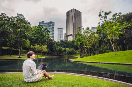 Man businessman or student in casual dress using laptop in a tropical park on the background of skyscrapers. Dressing in a white shirt, beige shorts. Young handsome businessman in casual clothes is sitting outside a business district. Mobile Office concepの写真素材