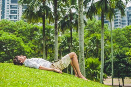 Man businessman or student in casual dress using laptop in a tropical park on the background of skyscrapers. Dressing in a white shirt, beige shorts. Young handsome businessman in casual clothes is sitting outside a business district. Mobile Office concepの写真素材