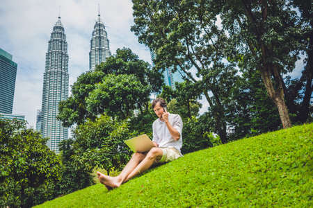 Man businessman or student in casual dress using laptop in a tropical park on the background of skyscrapers. Dressing in a white shirt, beige shorts. Young handsome businessman in casual clothes is sitting outside a business district. Mobile Office concepの写真素材