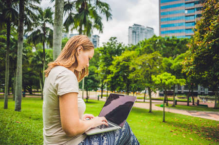 A young woman in casual dress using laptop in a tropical park on the background of skyscrapers. Mobile Office concept.の写真素材