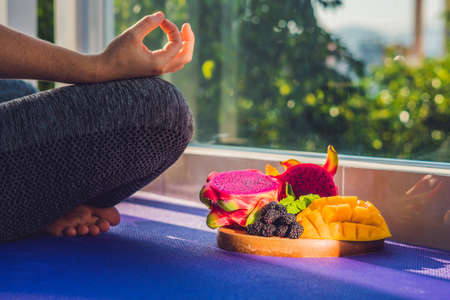 hand of a woman meditating in a yoga pose, sitting in lotus with fruits in front of her dragon fruit, mango and mulberry.の写真素材