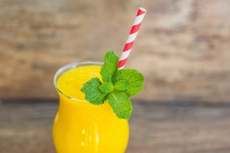 Juicy smoothie from mango in glass with striped red straw and with a mint leaf on old wooden background. Healthy life concept, copy space.の写真素材