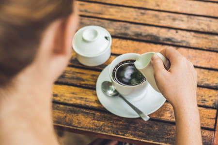 woman's hand pouring milk in white cup of coffee in outdoor cafe.の写真素材