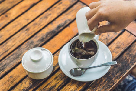 woman's hand pouring milk in white cup of coffee in outdoor cafe.の写真素材