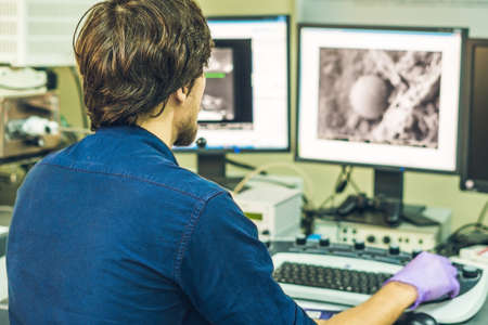 Scientist works at a electron microscope control pannel with two monitors in front of him.の写真素材
