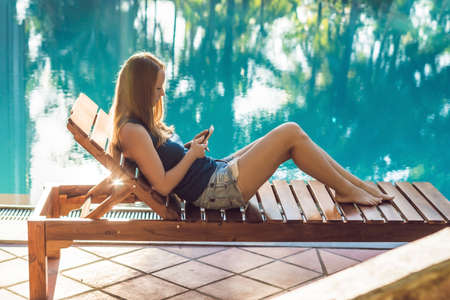 Happy smartphone woman relaxing near swimming pool. Beautiful girl using her mobile phone app 4g...