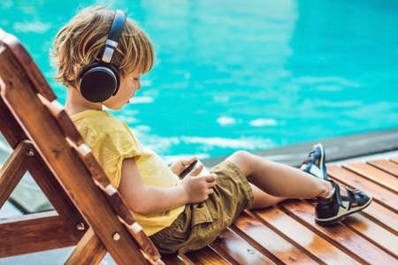 A cute little boy is using a smartphone and headphones lying on a deckchair by the pool. primary...