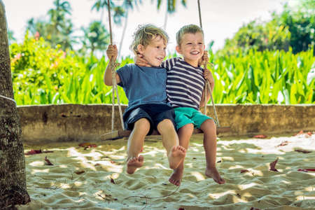 Two little blonde boys having fun on the swing on the tropical sandy coast.の写真素材