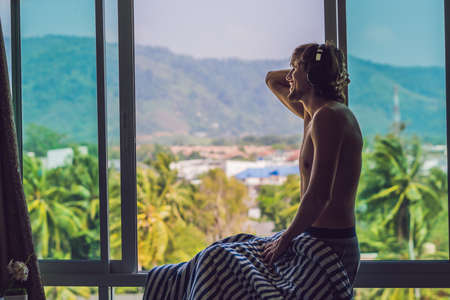 A man woke up in the morning and listens to music in wireless headphones on the background of a window overlooking the mountains.の写真素材