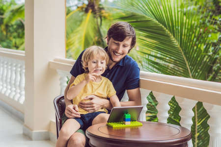 father and son in the tropics talking with friends and family on video call using a tablet and wireless headphones sitting on the terrace.の写真素材