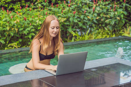 Young female freelancer sitting near the pool with her laptop in the hotel browsing in her smartphone. Busy at holidays. Distant work concept. Copy space for your text.の写真素材
