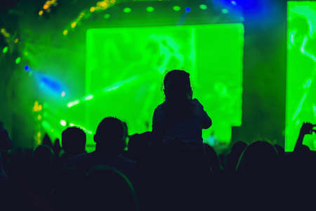 Silhouette of a big crowd at concert against a brightly lit stage. Night time rock concert with people having fun lifting hands up in the air and cheering the musicians.の写真素材