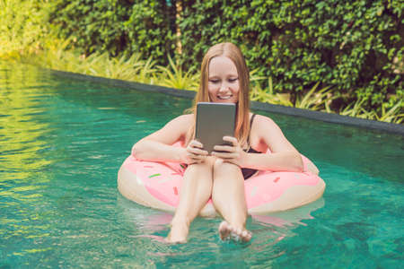 Young female freelancer sitting with a tablet in an inflatable circle in the pool. Busy at holidays. Distant work concept. Copy space for your text.の写真素材