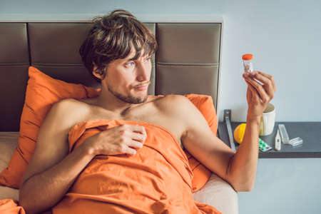 Man feeling cold, lying in the bed drinks tea and measures temperature with a thermometer.の写真素材