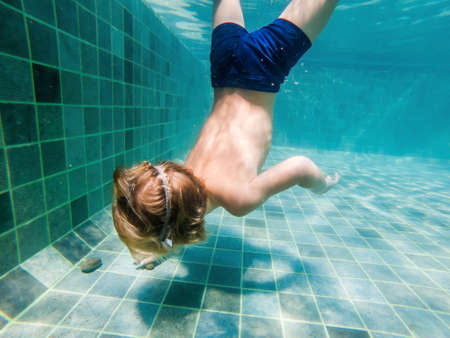 A child boy is swimming underwater in a pool, smiling and holding breath, with swimming glasses.の写真素材