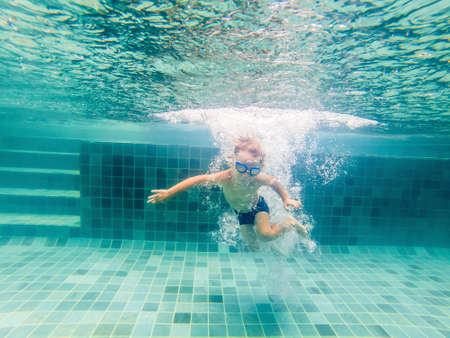 A child boy is swimming underwater in a pool, smiling and holding breath, with swimming glasses.の写真素材