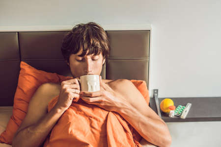 Man feeling cold, lying in the bed drinks tea and measures temperature with a thermometer.の写真素材