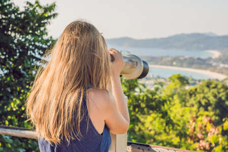 young beautiful blond woman enjoy the view with an coin operated binoculars. The water and the sky is blue. she wears a white dress and sunglasses. she feels good, is smiling and look to the horizon.の写真素材