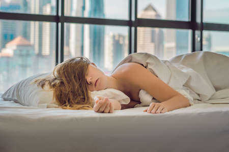 Woman wakes up in the morning in an apartment in the downtown area with a view of the skyscrapers. Life in the noise of the big city concept. Not enough sleep.の写真素材