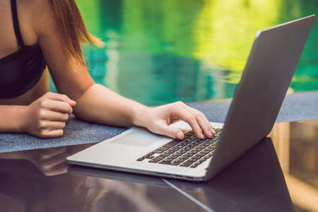 Young female freelancer sitting near the pool with her laptop in the hotel browsing in her...