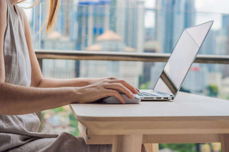 Young woman is working on a laptop on her balcony overlooking the skyscrapers. Freelancer, remote...