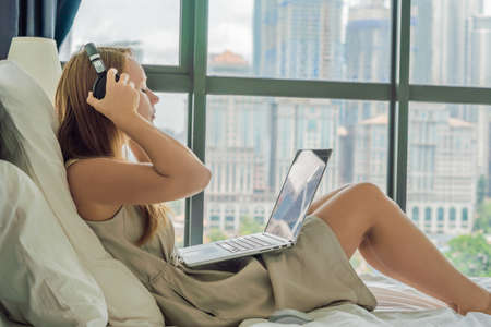 Young woman is working on a laptop in his bed on a background of a panoramic window overlooking the skyscrapers.の写真素材