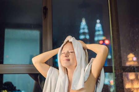 Beautiful woman wipes her hair after a shower on the background of a window with a panoramic view of the night city.の写真素材