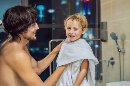 Dad wipes his son with a towel after a shower in the evening before going to sleep on the background of a window with a panoramic view of the city.の写真素材