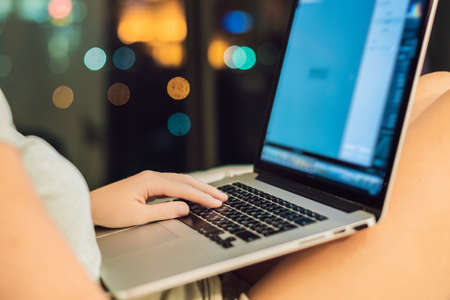 young beautiful, caucasian, red haired, internet addicted woman working bored, sleepless and tired on her laptop in bed in her bedroom late at night in dark room on the background of a window overlooking the big city. looking annoyed.の写真素材