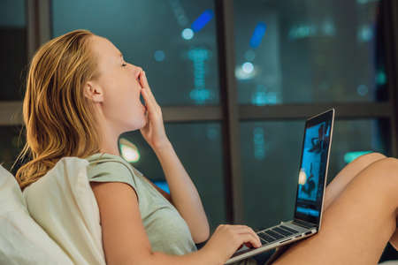 young beautiful, caucasian, red haired, internet addicted woman working bored, sleepless and tired on her laptop in bed in her bedroom late at night in dark room on the background of a window overlooking the big city.の写真素材